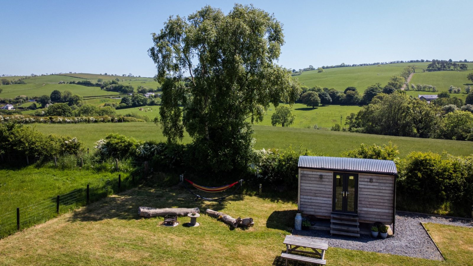 Cae Main: A cozy wooden cabin with a bench and fire pit, nestled in a grassy field beneath a clear sky.