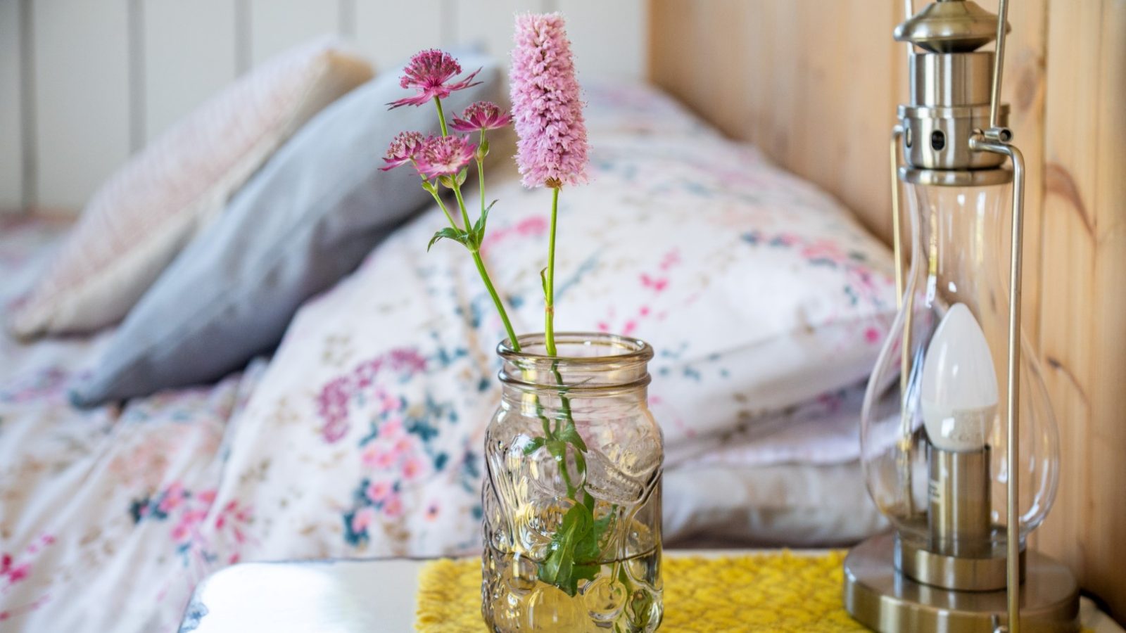 A glass jar with pink flowers sits on a yellow mat in a charming shepherds hut, beside a bed with floral sheets.