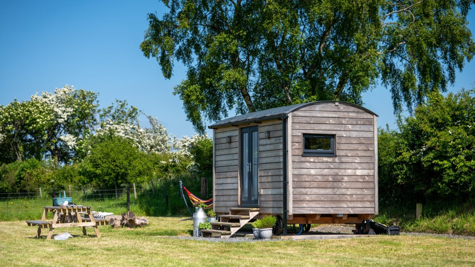 Cae Main wooden tiny house with a front porch, in a grassy field surrounded by trees, with a picnic table and hammock.