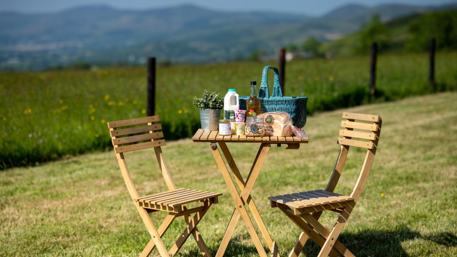 Two wooden chairs and a table with bread, milk, and drinks near the Shepherds Hut on a grassy field with hills.