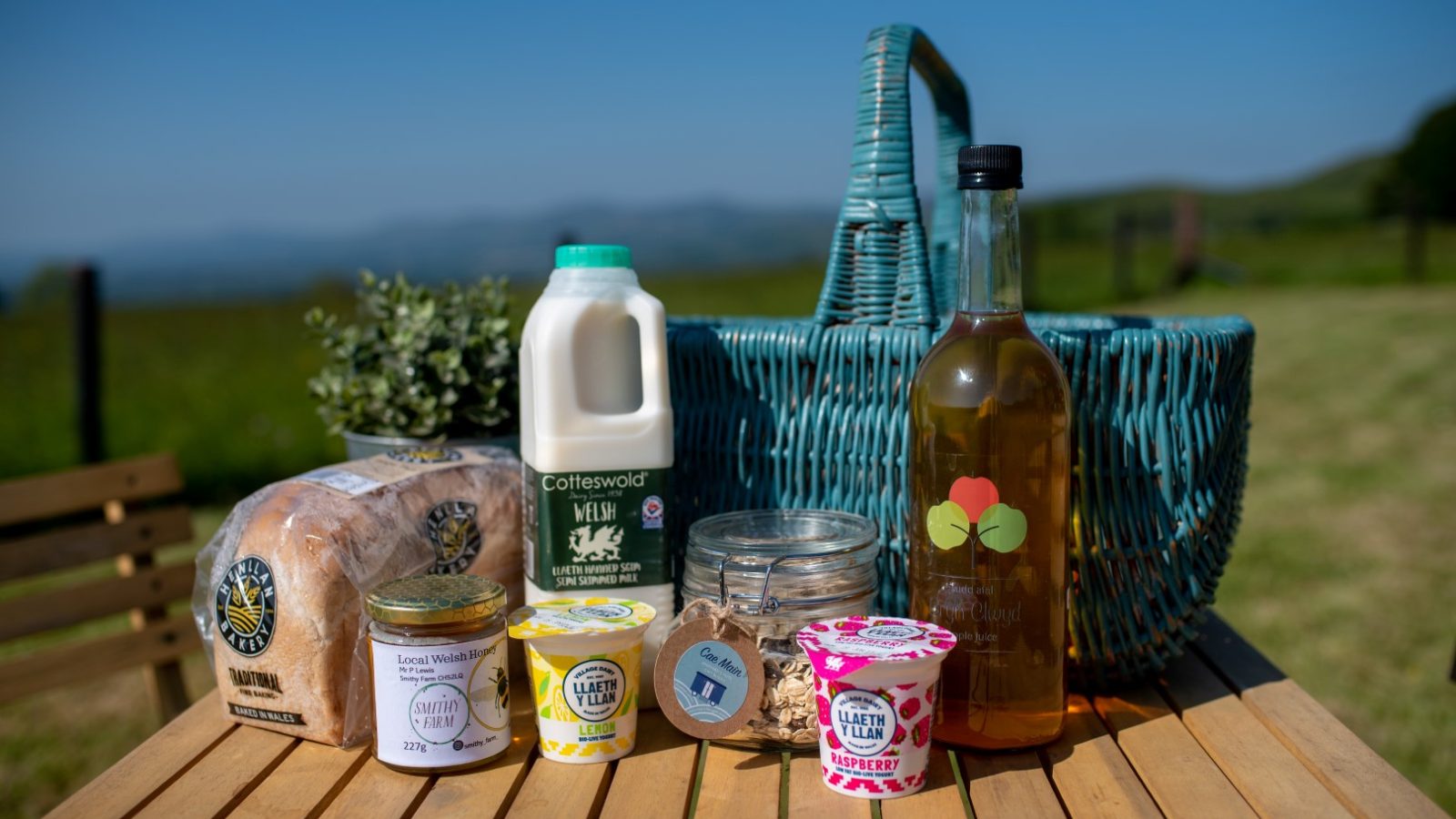 A picnic table with bread, milk, jars, juice, and yogurt near a wicker basket by Cae Main's grassy landscape.