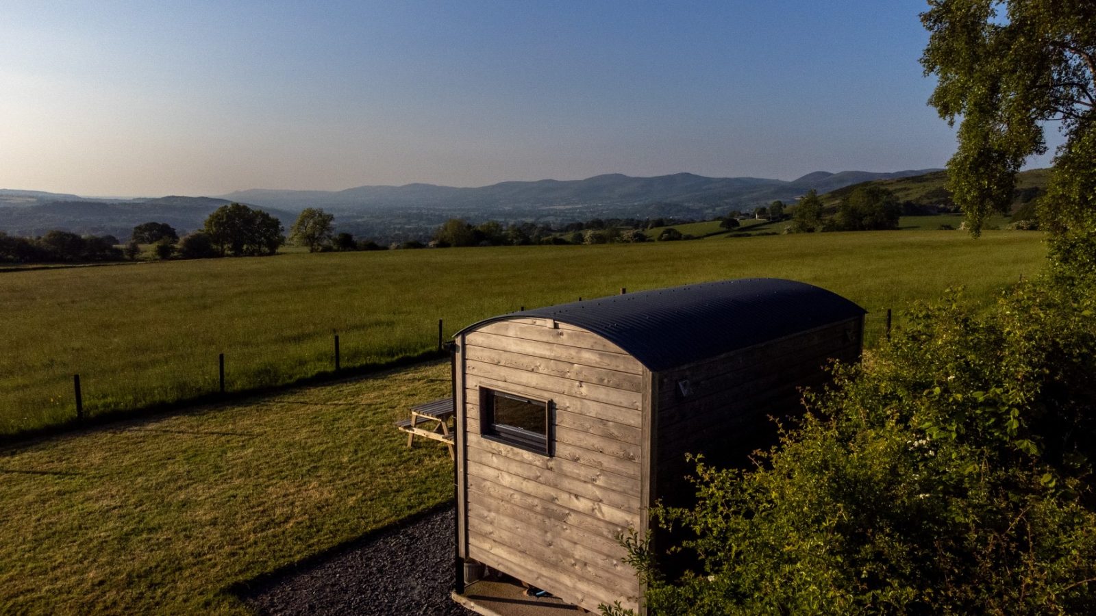 Shepherds Hut on a grassy field with distant mountain range under a clear blue sky near Cae Main.