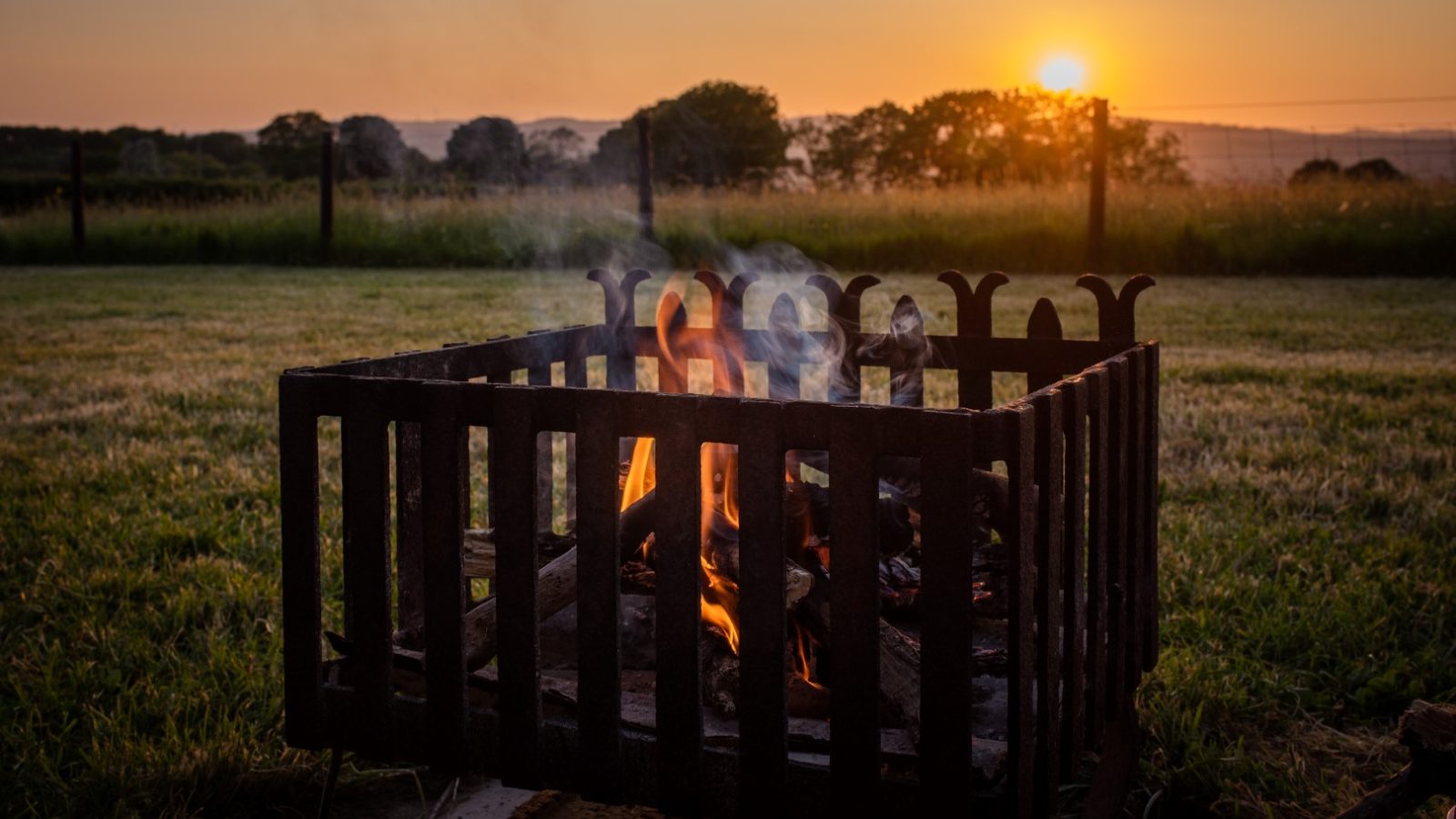 A metal fire pit with crackling wood emits smoke by the Shepherds Hut, set in a grassy field at sunset near Cae Main.