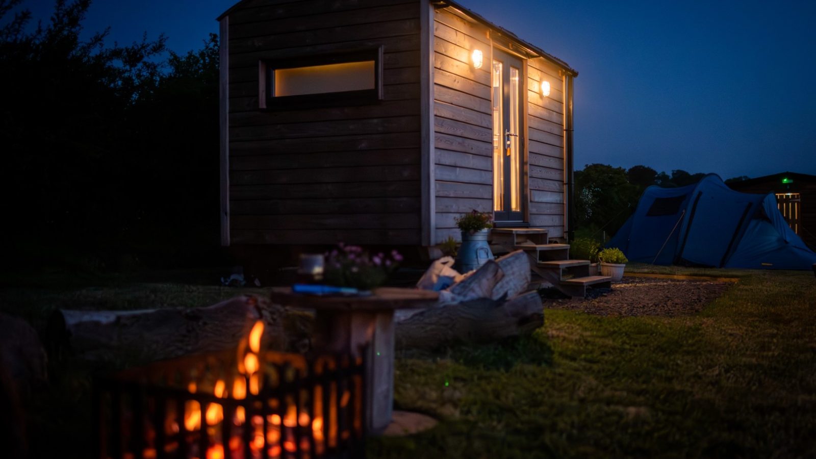 A small wooden cabin and Shepherds Hut with lights on, next to a campfire and tent, under a dusky blue sky in a grassy area.