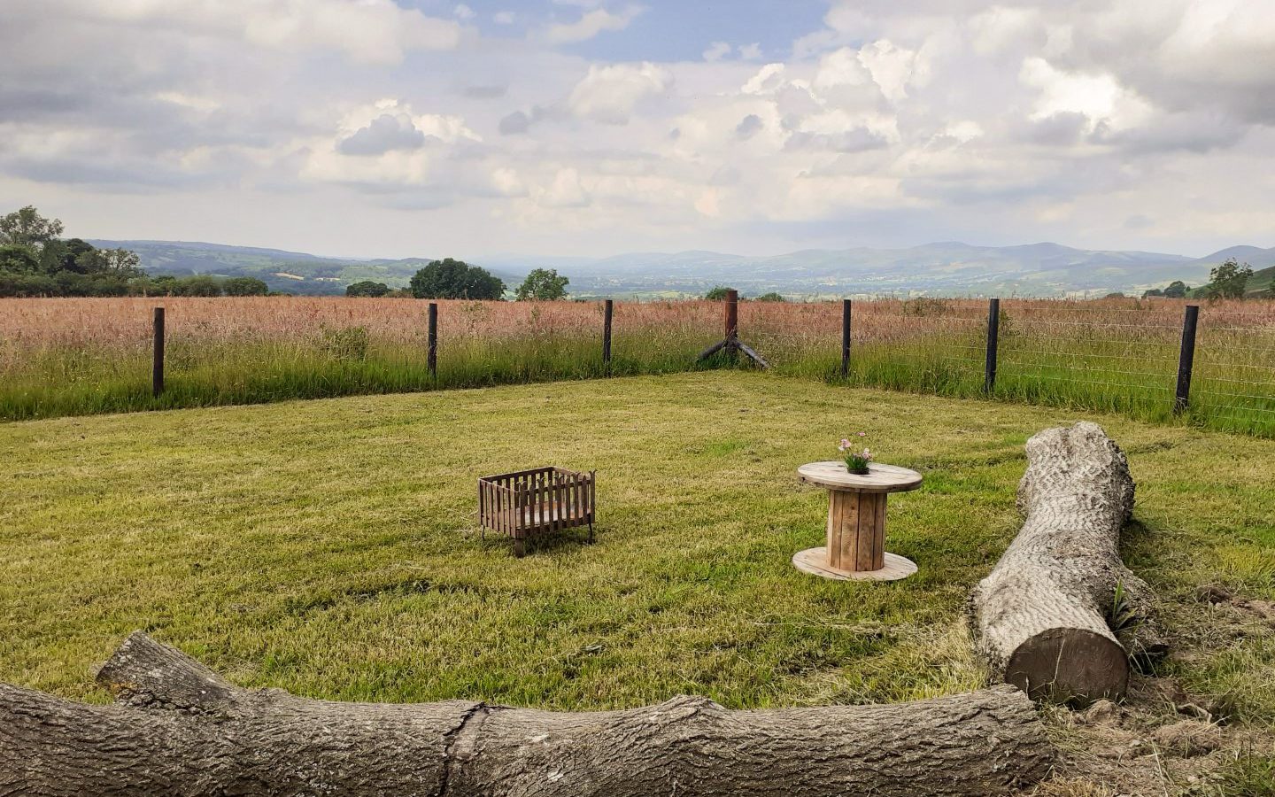 A peaceful rural scene at Cae Main Shepherds Hut features a grassy area with a wooden spool table holding a small vase. A log lies in the foreground, and a wire fence surrounds the field. In the background, rolling hills stretch under a partly cloudy sky, evoking the charm of an inviting shepherd's hut setting.