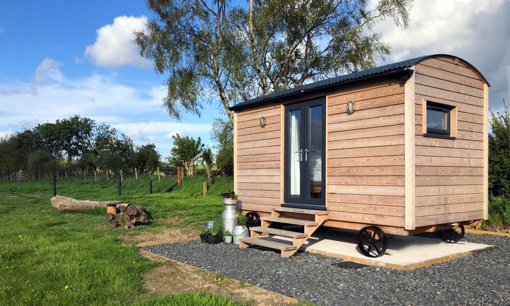 Nestled on a grassy field, this charming wooden shepherd's hut sits peacefully with logs nearby and a tree in the background. The quaint Cae Main setting adds to its rustic allure, perfect for a serene escape.