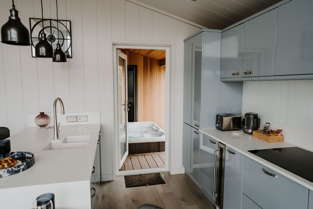 The modern kitchen in Bowland Retreat features white countertops and light blue cabinets. A white sink and stainless steel fixtures are visible. Through an open door, a wooden deck with a hot tub is seen, while hanging pendant lights enhance the contemporary feel. Perfect for cozy lodges!