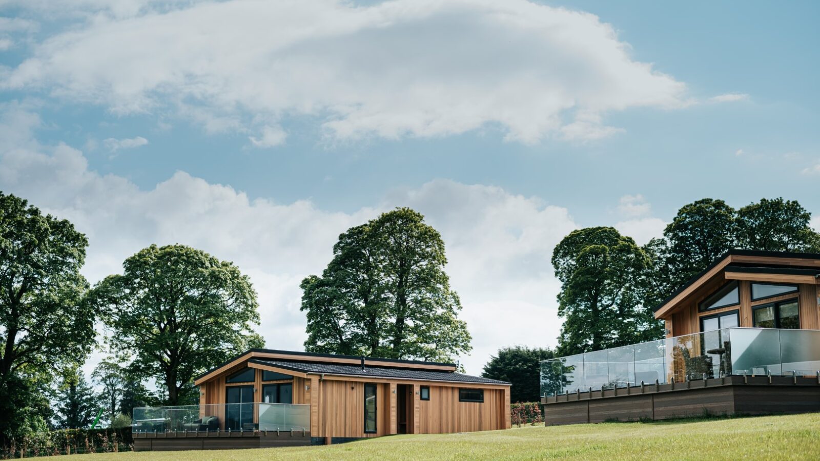 Two modern wooden lodges with large windows and glass railings are situated on a grassy hillside, surrounded by trees and a blue sky, embodying the serene charm of Bowland Retreat Lodges.