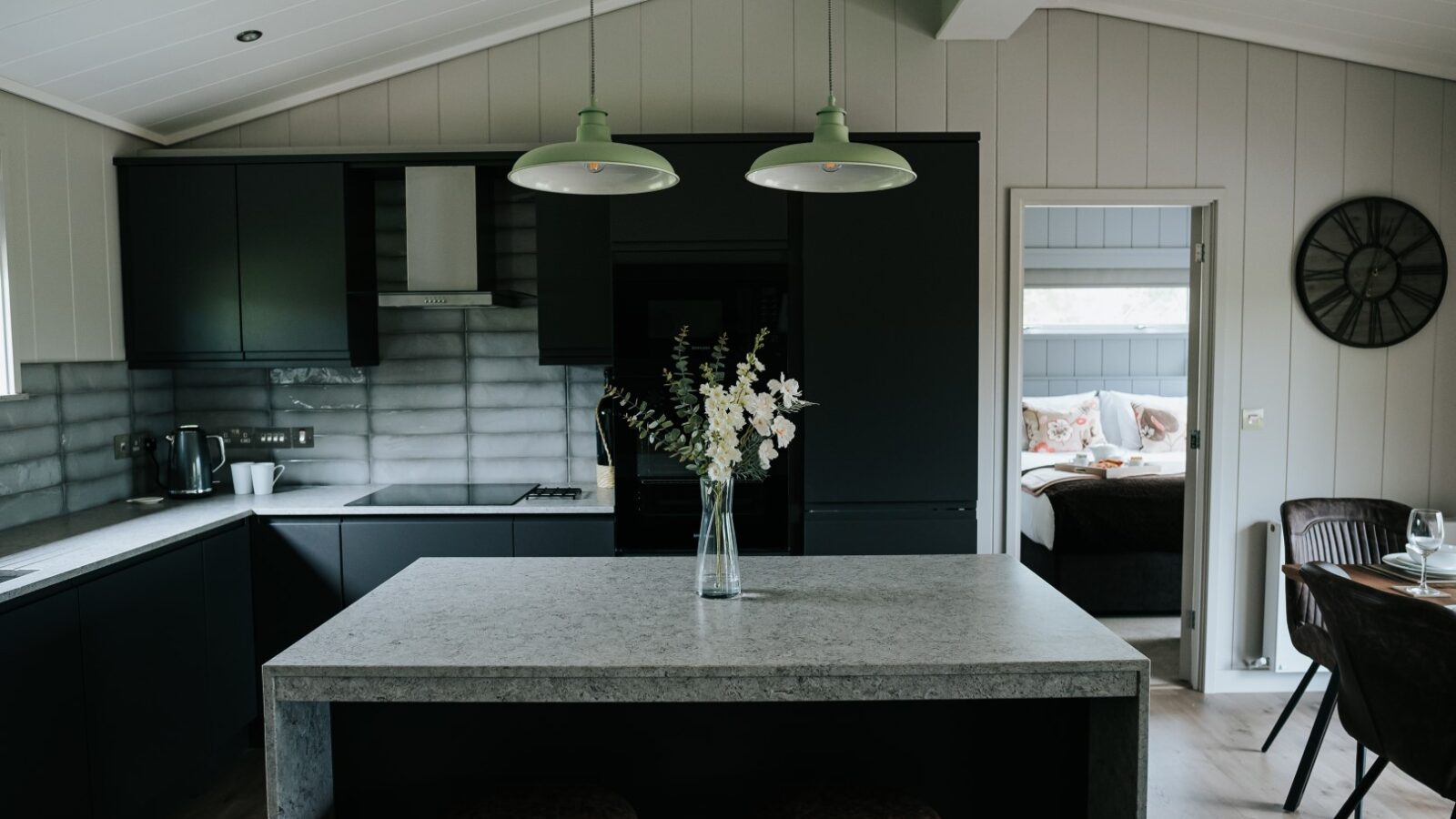 Modern kitchen with dark cabinetry, large island adorned with a vase of flowers, and pendant lights enhancing the ambiance. Through an open door, the bedroom welcomes you. Perfect for those seeking a contemporary escape at Bowland Retreat Lodges.