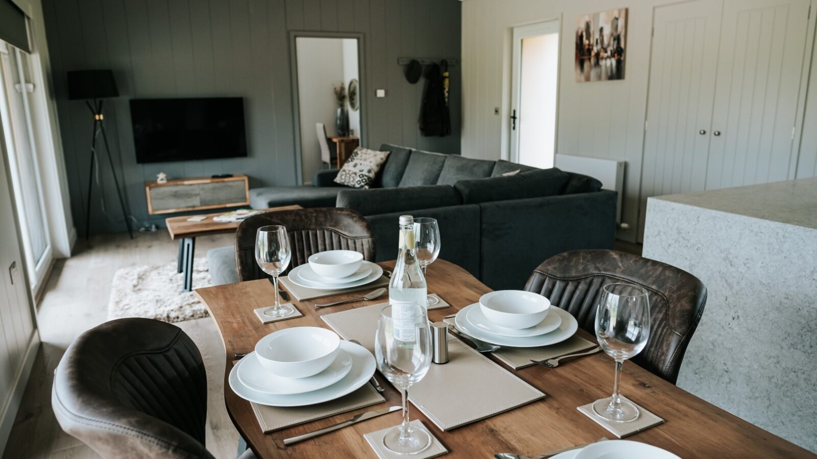 The Bowland Retreat offers a modern living room with gray walls, where a flat-screen TV, a comfortable L-shaped sofa, and a dining table set elegantly with white dishes and glasses create the perfect lodge atmosphere.