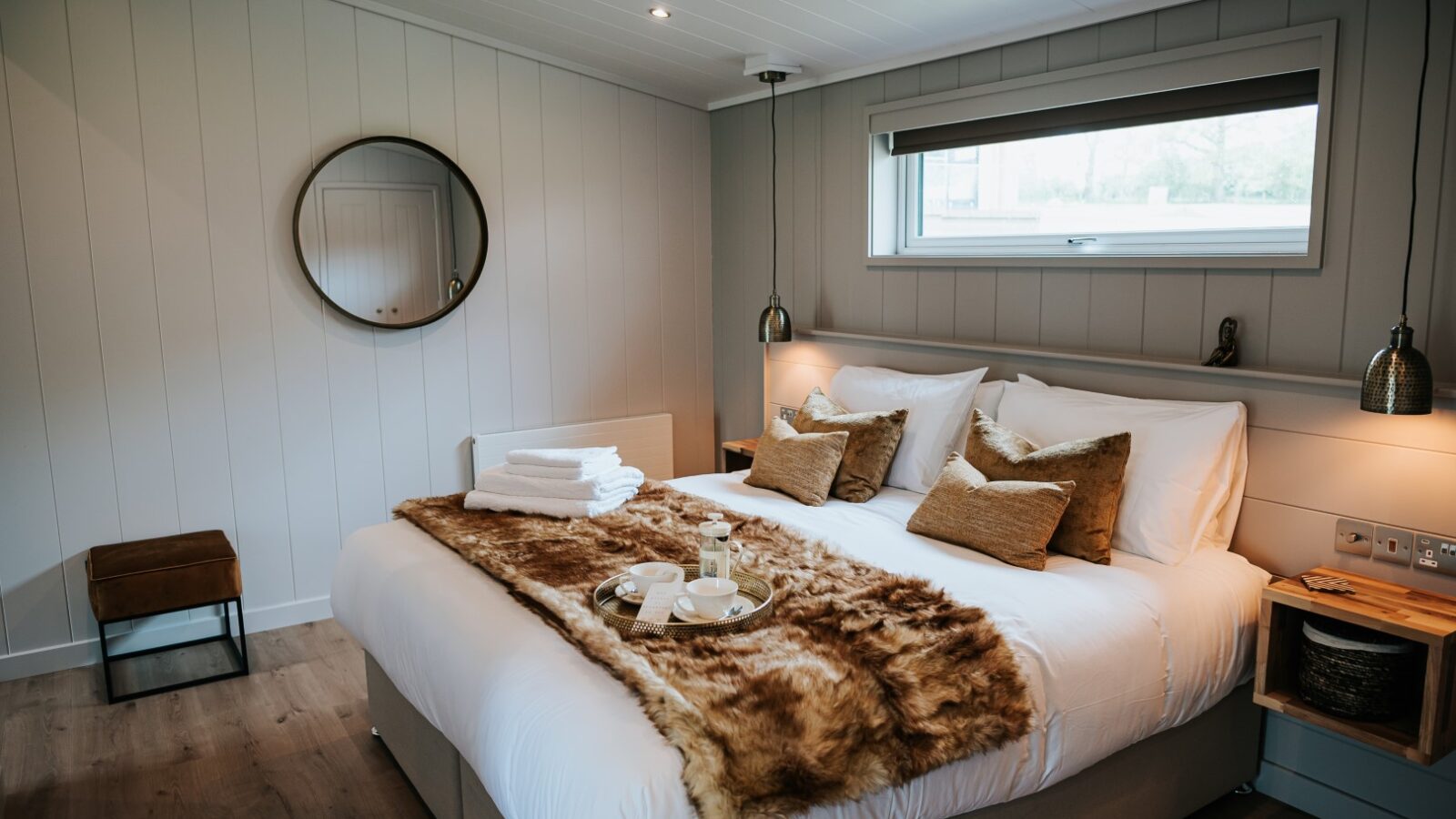 Experience the charm of Bowland Retreat in this cozy bedroom featuring a double bed adorned with a fur throw, four plush pillows, and soft bedside lamps. A small window invites natural light, while a wall mirror and wooden stool complete this inviting lodge sanctuary.