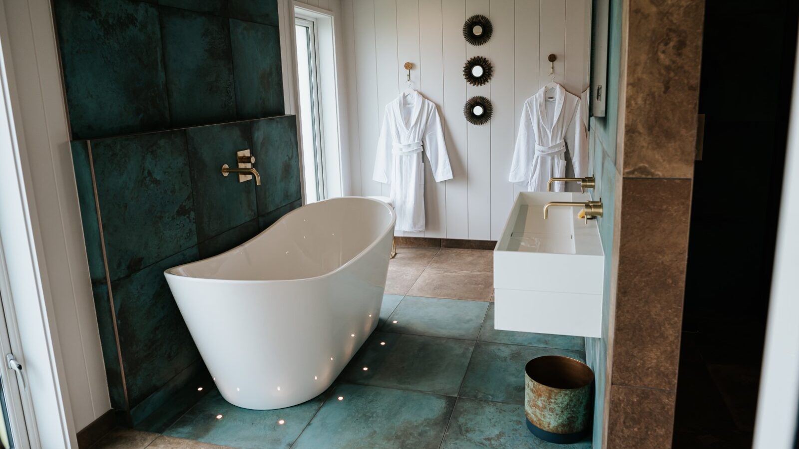 Experience a modern bathroom retreat featuring a freestanding tub, two hanging white robes, a double sink, and decorative wall accents. Perfectly designed to provide relaxation akin to the serene surroundings of Bowland Retreat Lodges.