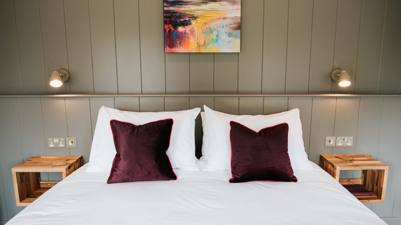 A neatly made bed with purple pillows, bedside tables, and wall lights sits perfectly against a gray paneled wall beneath an abstract painting, offering a serene vibe reminiscent of Bowland Retreat's elegant lodges.