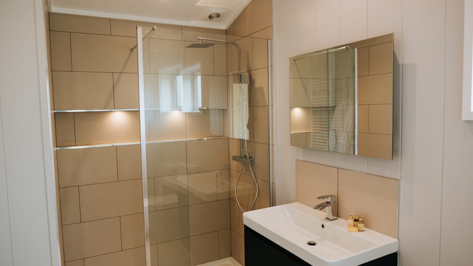 Modern bathroom at Bowland Retreat features a large beige-tiled shower, glass door, sleek vanity with white sink, and mirrored cabinet, offering a tranquil escape that perfectly complements the rustic charm of lodges.