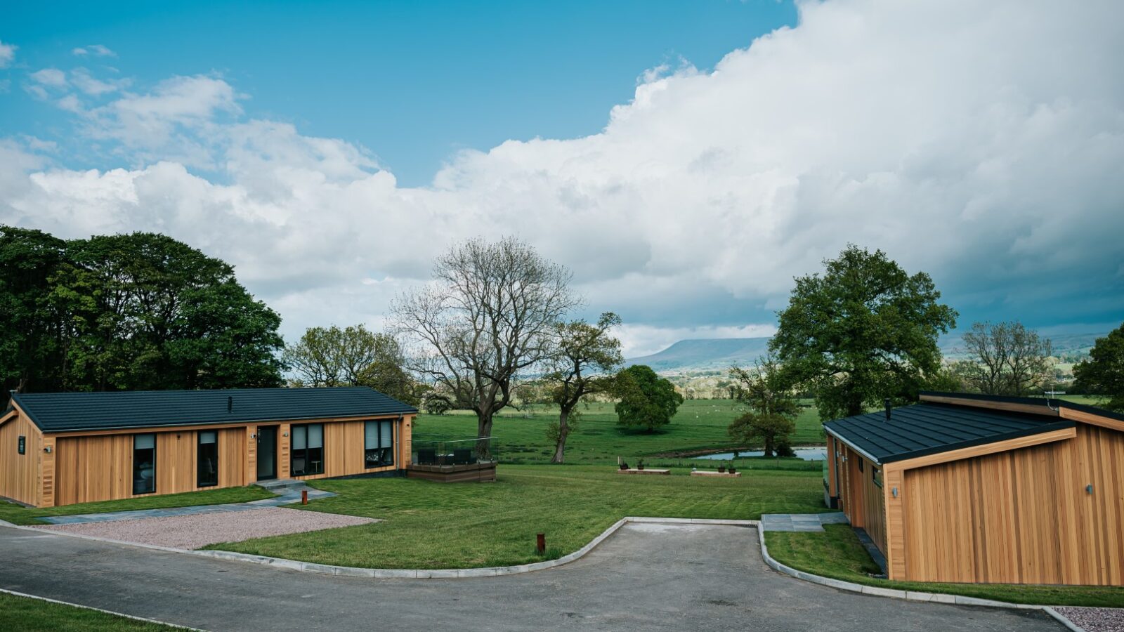 Nestled in a rural landscape, the modern wooden cabins of Bowland Retreat are surrounded by lush greenery and trees under a cloudy sky.