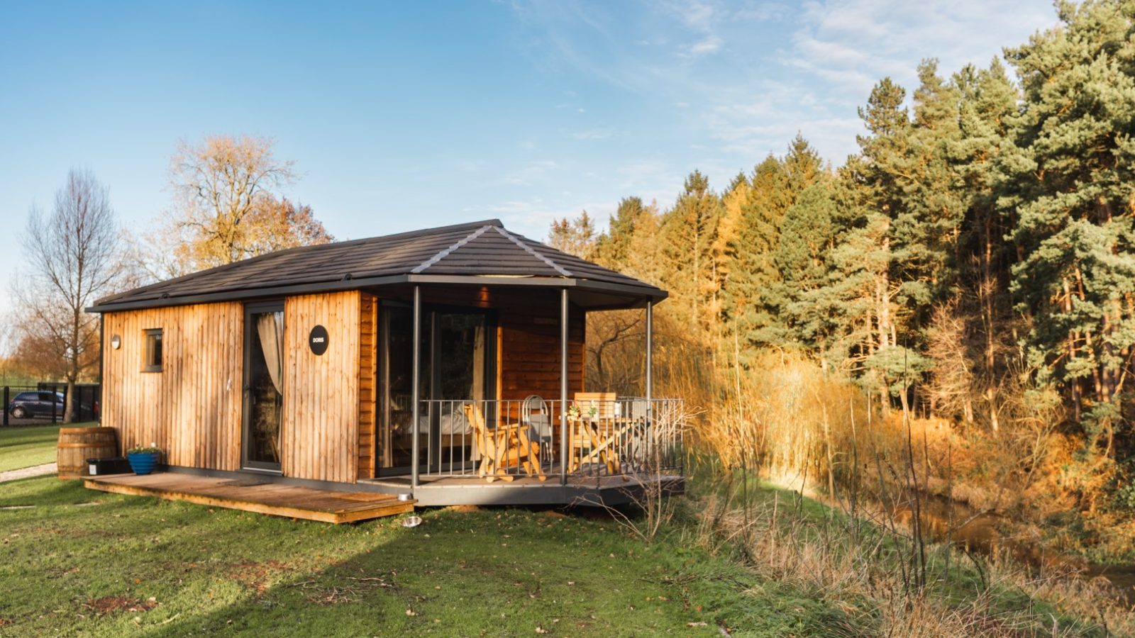 A quaint wooden cabin with a slanted roof and a porch overlooks the riverside. Chairs rest on the porch, inviting relaxation. Surrounded by trees adorned with autumn foliage, the cabin basks under a clear blue sky, promising a sunny day. Riverside Cabins Shropshire