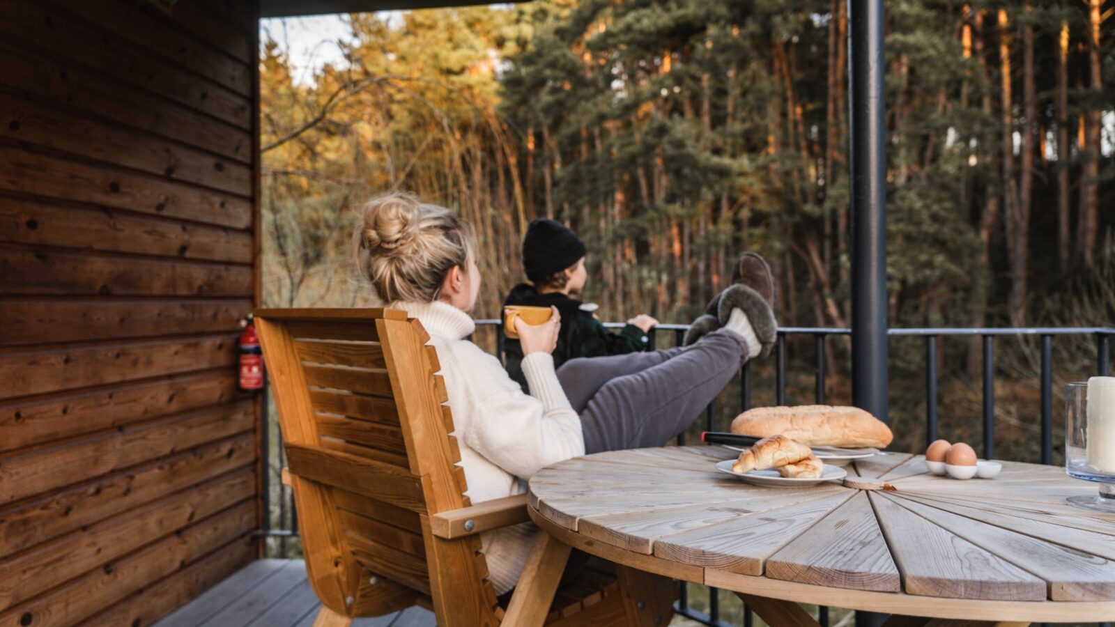 Two people unwind on a wooden deck of a riverside cabin, with bread and eggs resting on the table, surrounded by trees.