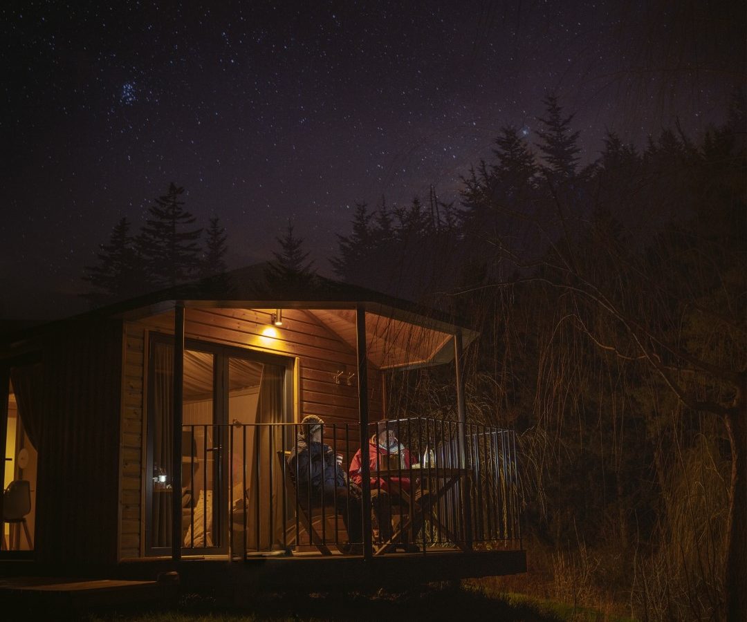 A cosy riverside cabin with a glowing light on the porch is set against a backdrop of a starry night sky. Tall trees surround the cabin, and two chairs rest on the porch, creating a peaceful nighttime scene by the gentle flow of water.