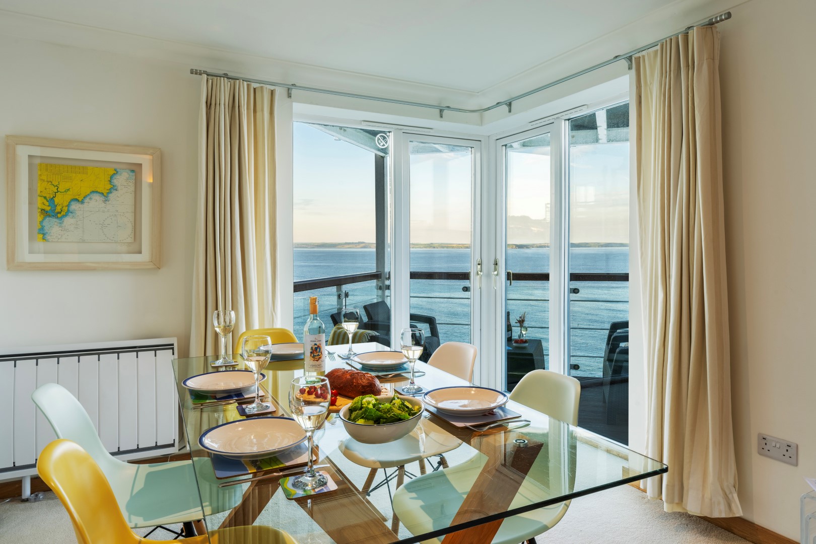 In a cozy September UK staycation setting, enjoy a dining table elegantly set with plates and wine on a glass-top surface by the window, offering stunning ocean views amidst vibrant yellow chairs.