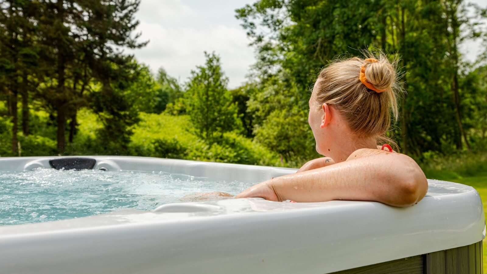 A person unwinds in an outdoor hot tub by a cozy riverside cabin, surrounded by lush greenery and trees on a sunny day.