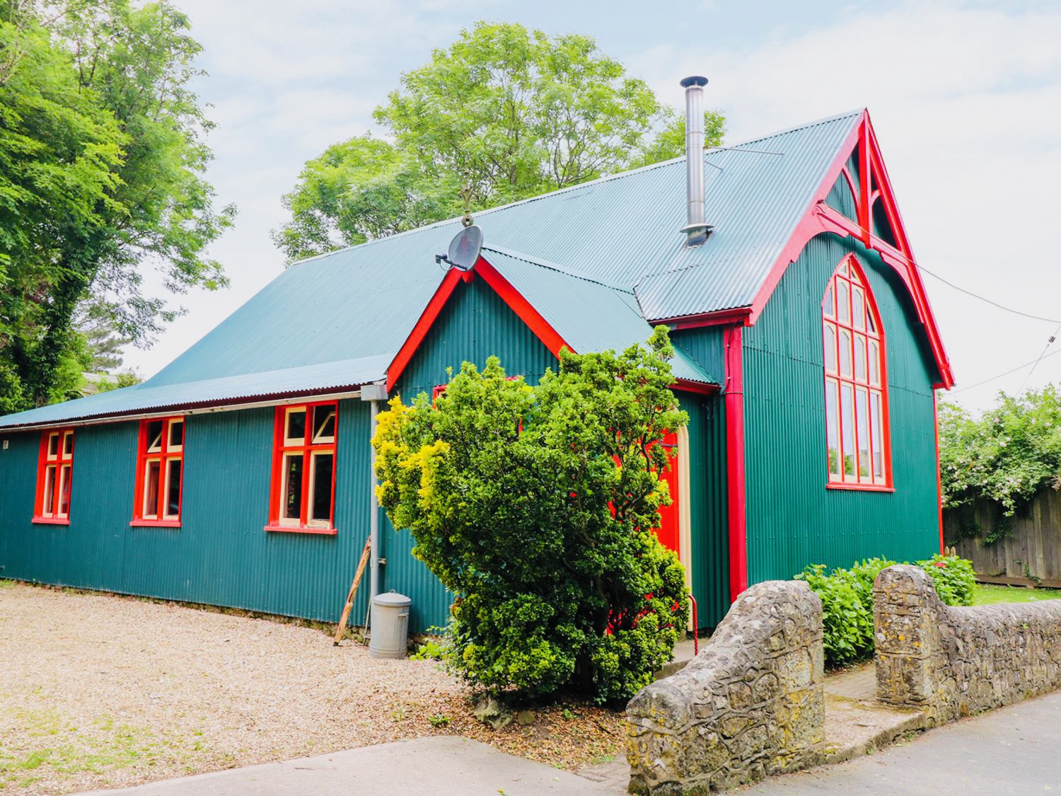 A vibrant green and red house with a corrugated metal roof, perfect for a September UK staycation, is surrounded by lush greenery and a charming stone wall.