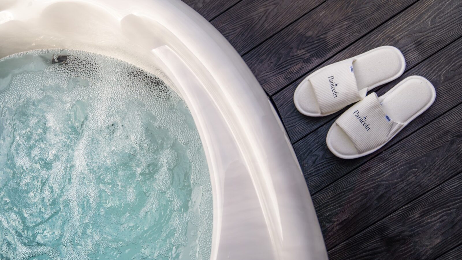 A bubbling hot tub sits on a dark wooden deck, accompanied by a pair of white slippers labeled 