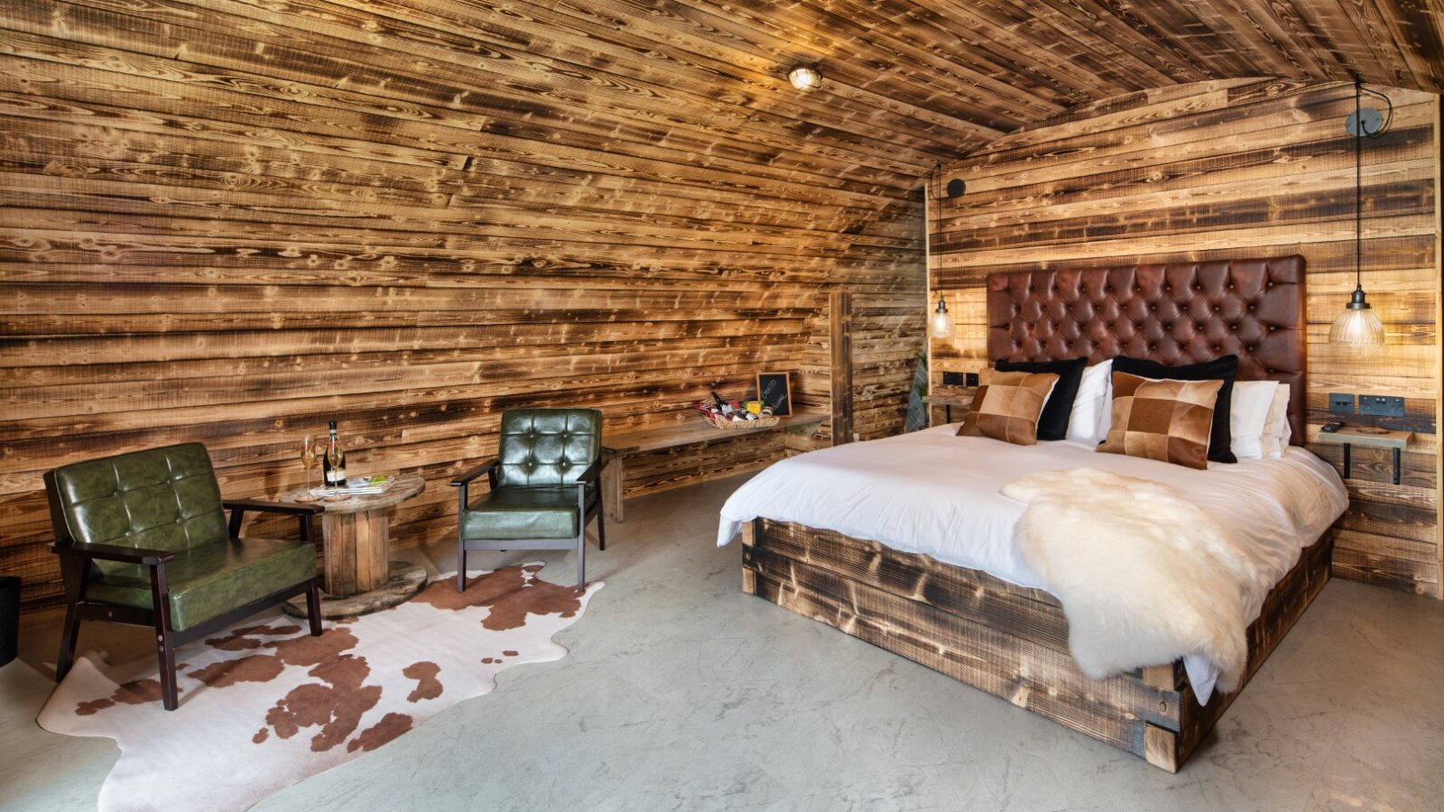 Rustic bedroom featuring wooden walls and a large bed, complemented by green chairs and a cowhide rug on the concrete floor. The space exudes a cozy, natural ambiance akin to a Piglet Pod retreat.