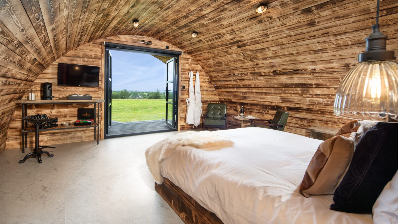 Cozy wood-paneled Piglet Pod with a double bed, modern lamp, wall-mounted TV, and open doors revealing a grassy field view.