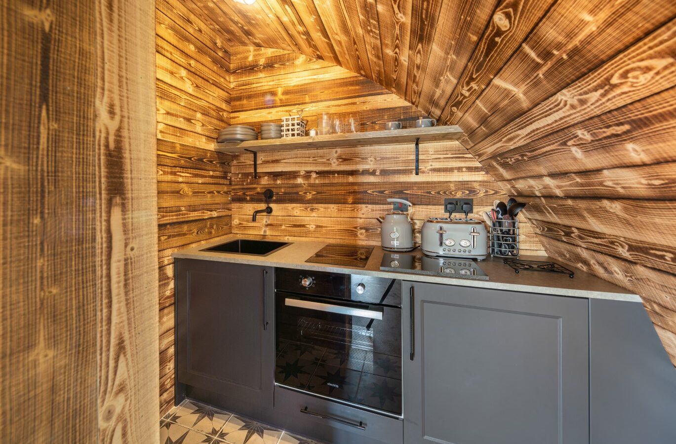 The charming Piglet Pod-inspired wooden kitchen boasts a sloped ceiling, complete with a cozy sink, stove, sleek grey cabinets, and a stylish patterned tile floor.