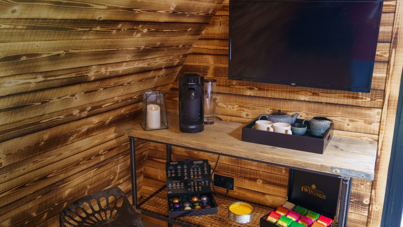 A cozy nook featuring a Piglet Pod alongside a wall-mounted TV, coffee maker, teacups, and assorted tea and coffee pods on a rustic wooden table.