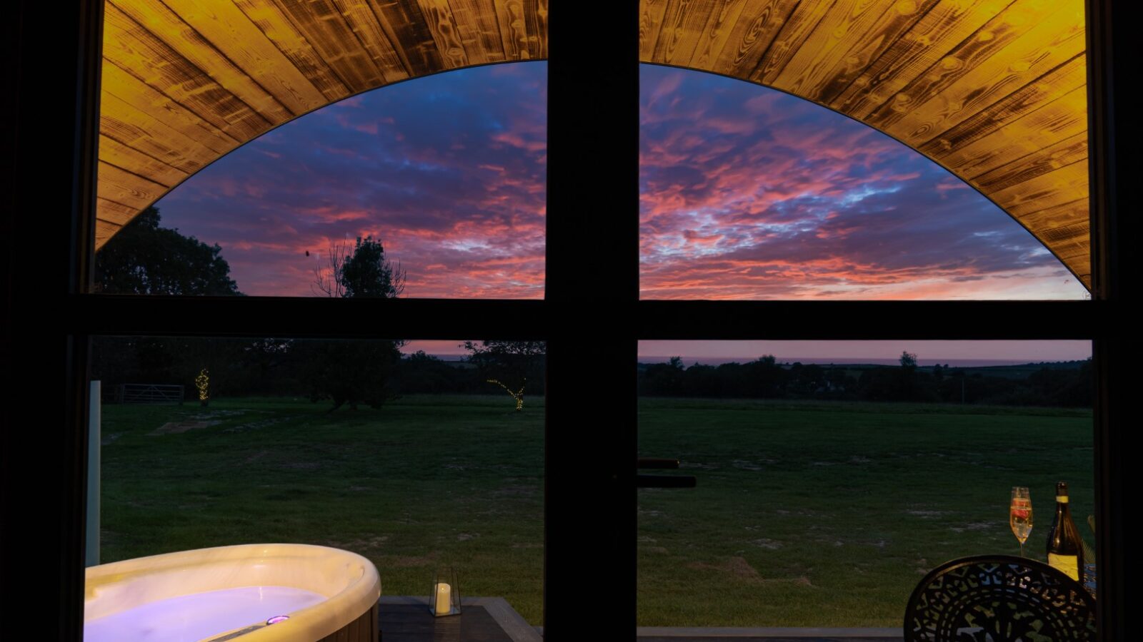 View through the Piglet Pod's window of a patio with a hot tub, trees, and vibrant sunset sky. Two champagne glasses rest on a small table.