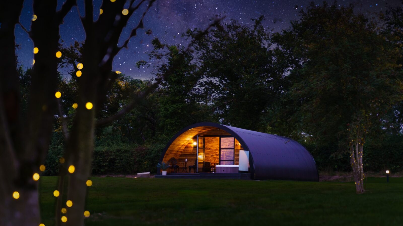 A small modern Piglet Pod cabin with a curved roof is illuminated at night, surrounded by trees and fairy lights under a starry sky.