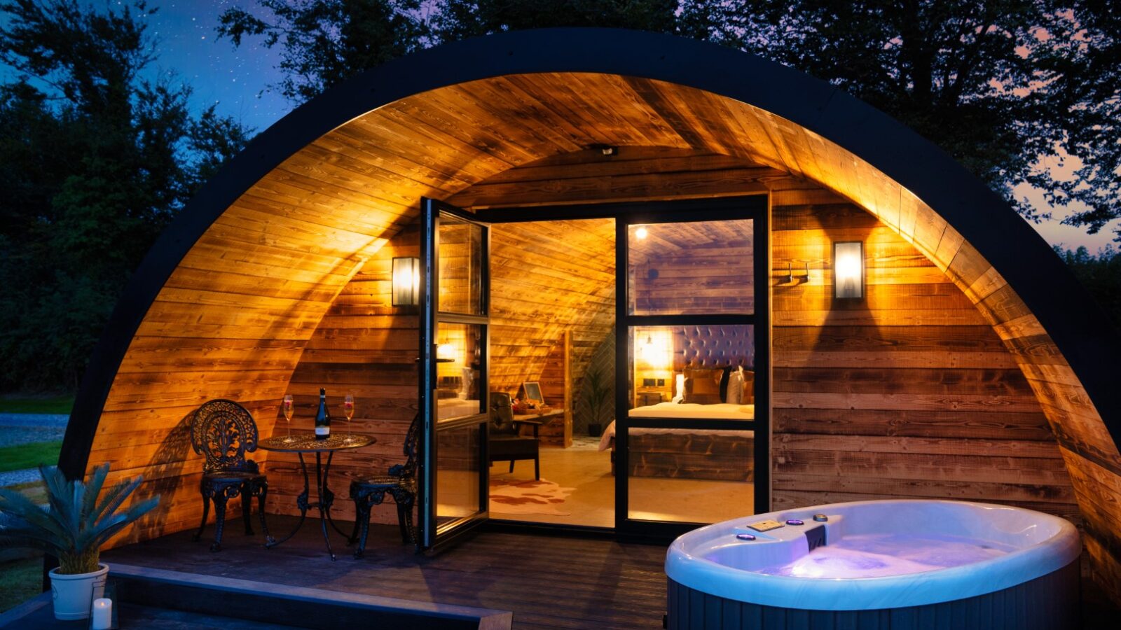 Discover the enchanting Piglet Pod: a wooden cabin with open doors, cozy patio seating, a soothing hot tub, and warm lighting embraced by dusk. All set against a backdrop of trees and a starry sky for an unforgettable retreat.