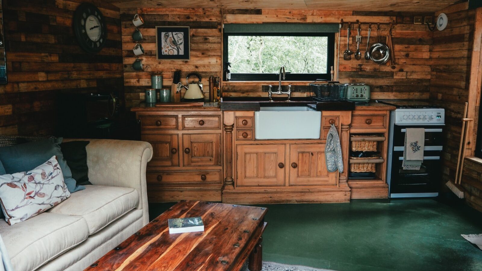 Beneath the Branches, this rustic wooden kitchen and living area features a cozy sofa, a charming coffee table, a farmhouse sink, and a traditional stove with kitchenware neatly displayed on open shelves.