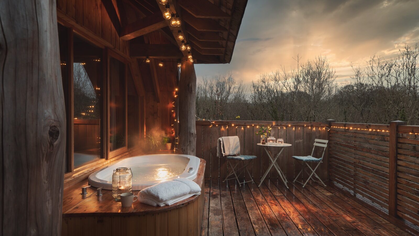 The outdoor hot tub on a wooden deck at Wolf Wood Treehouses is perfectly adorned with string lights, plush towels, and a small table set. Surrounded by trees and overlooking a stunning sunset, it offers the ultimate tranquil escape.