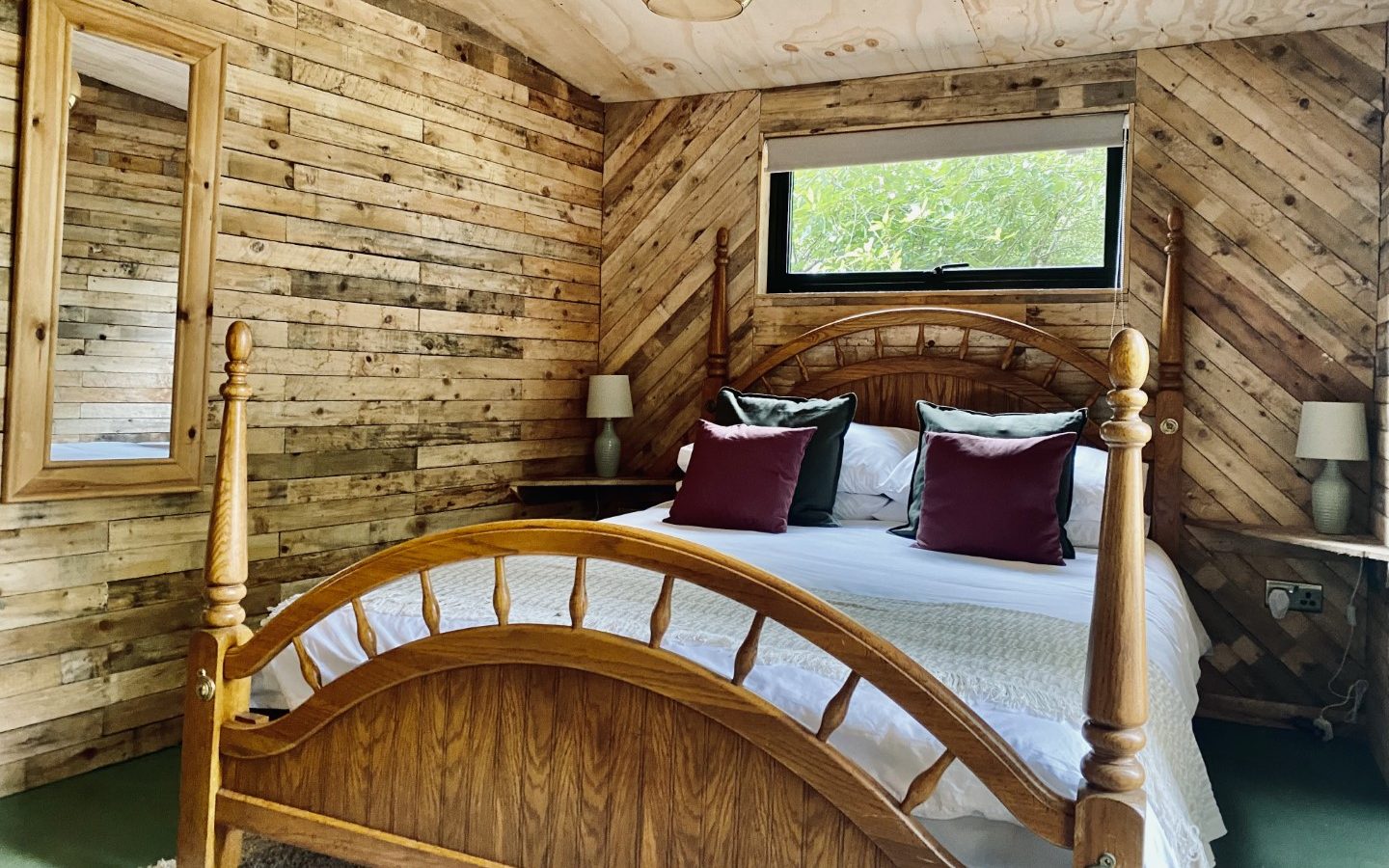 Beneath the Branches, a cozy bedroom features wooden walls and a sturdy wooden bed frame. Two soft pillows rest beneath a window, framed by the gentle glow of lamps on twin bedside tables.