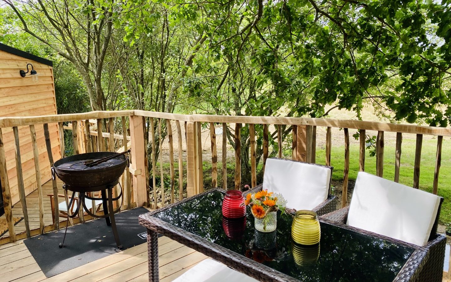 Beneath the branches, a wooden deck hosts a black grill and a glass table set for two. The table is adorned with a bouquet of flowers and two colored lanterns. Green leafy trees envelop the space, creating a peaceful and natural setting.