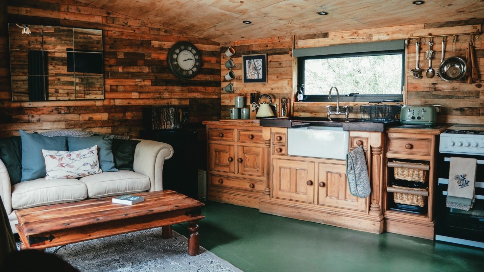 Beneath the Branches, this cozy, rustic living room and kitchen blend wooden accents with charm. A beige sofa invites relaxation by a wooden coffee table, while a farmhouse sink sits beneath a window. Kitchen utensils hang above, completing the scene adorned with decorations and a timeless clock.