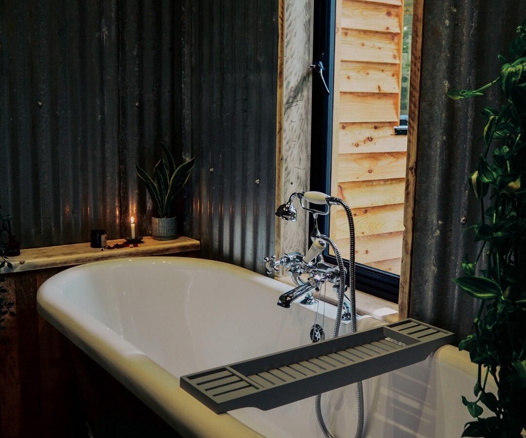 A white clawfoot bathtub sits beneath the branches of an inviting bathroom setting, framed by corrugated metal walls. A window casts natural light onto subtle accents like candles and a potted plant on the ledge, creating a serene retreat.