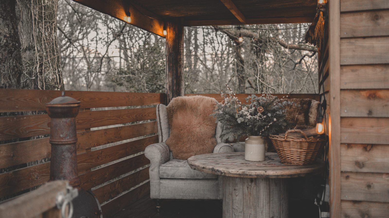 Cozy outdoor seating area nestled beneath the branches, with a cushioned armchair draped in fur beside a rustic wooden table. The table holds a wicker basket and floral arrangement. Wooden railings and warm string lights envelop the space, surrounded by bare trees.