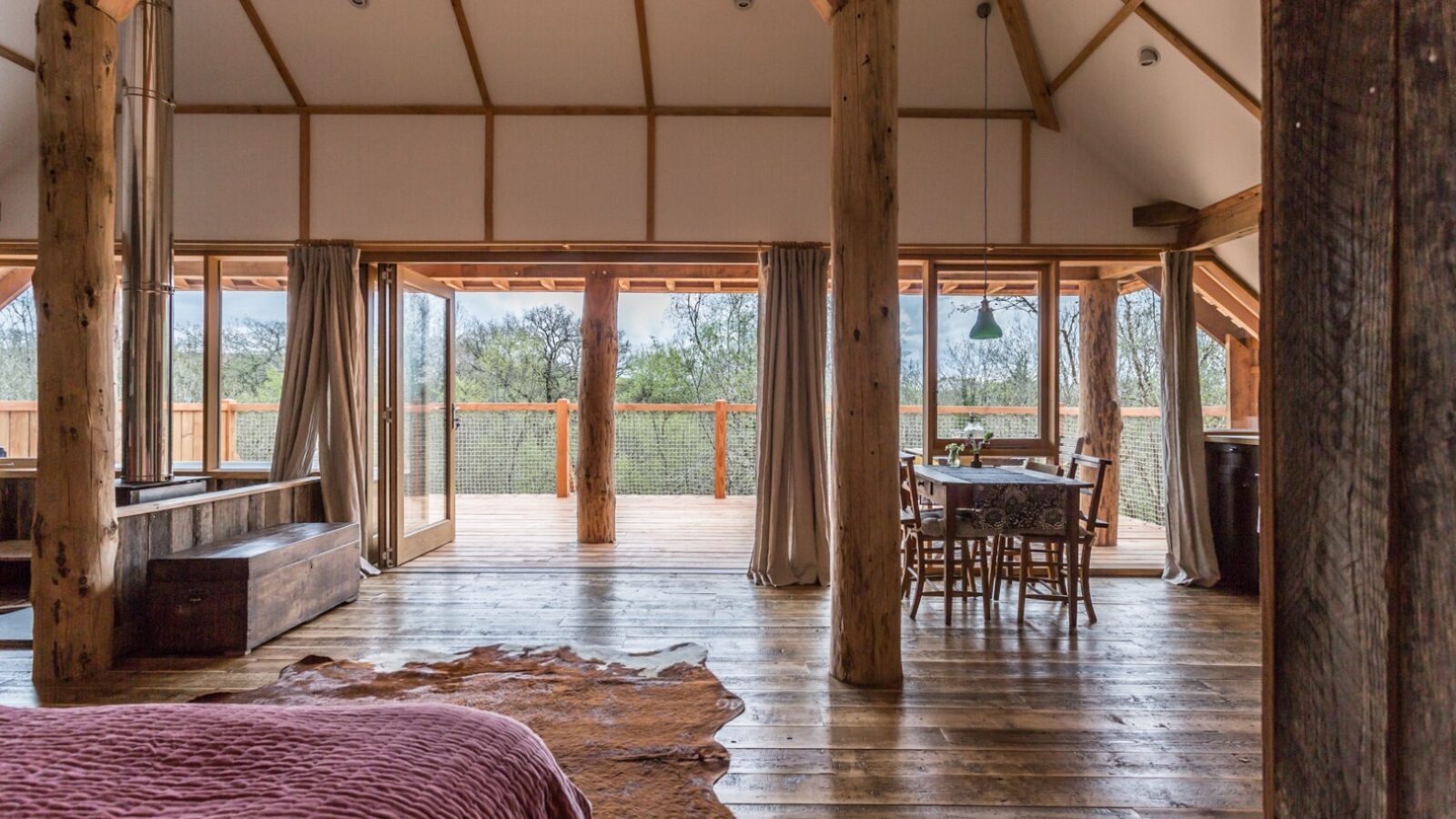 Spacious rustic cabin interior in Wolf Wood with large windows, wooden beams, dining table, and bed. This charming retreat opens to a deck that offers an outdoor view reminiscent of enchanting treehouses.