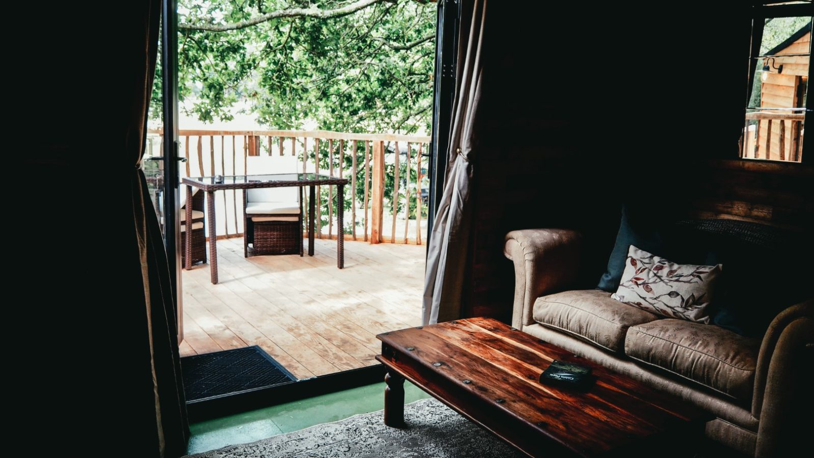 Cozy interior of a room featuring a beige sofa and wooden coffee table, leading to an outdoor deck with a table and chairs. Beneath the branches of a lush, green tree, the deck creates a serene and inviting atmosphere.