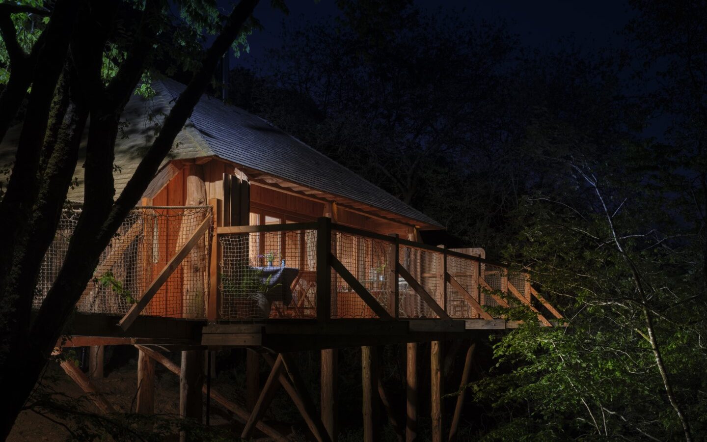 A treehouse at night, partially illuminated, rests elegantly in the heart of Wolf Wood Treehouses. Its wooden deck, enveloped by a serene forest canopy, offers a tranquil escape surrounded by nature's whispers.