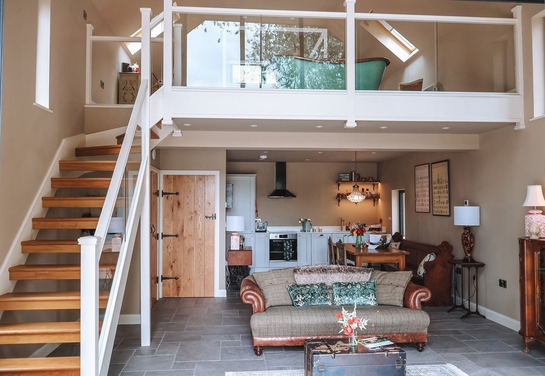 A cozy two-story living space reminiscent of a charming Boat House, with a wooden staircase leading to a mezzanine. The lower level features a couch, coffee table, and kitchen area with modern appliances. The decor includes plants, a table lamp, and framed art on the walls.