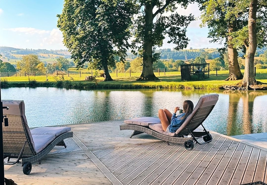 A person relaxes on a lounge chair by the serene lakeside at The Boat House, surrounded by large trees and rolling green hills under a blue sky. The tranquil setting features two lounge chairs on a wooden deck overlooking the lake.