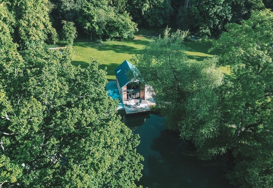 A secluded, modern cabin with a triangular roof sits beside a tranquil pond, resembling a serene boathouse setting. Surrounded by lush green trees and a grassy clearing, sunlight filters through the foliage, enhancing the idyllic atmosphere.