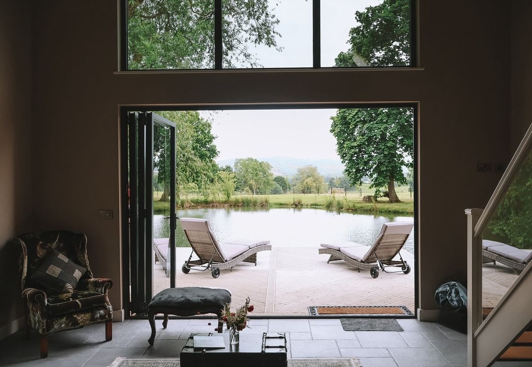 Luxurious room with open glass doors leading to a wooden deck overlooking a serene pond. Two lounge chairs are on the deck, surrounded by lush greenery and distant hills under a cloudy sky. Enjoy waterfront dining or try boat rentals for an enhanced experience before settling into comfortable seating inside.