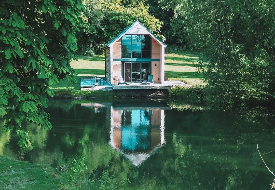 A modern, glass-fronted Boat House surrounded by lush greenery is reflected in a serene pond, offering a picturesque setting with a grassy lawn and trees. This peaceful, natural atmosphere makes it ideal for lakeside restaurant experiences or waterfront dining.