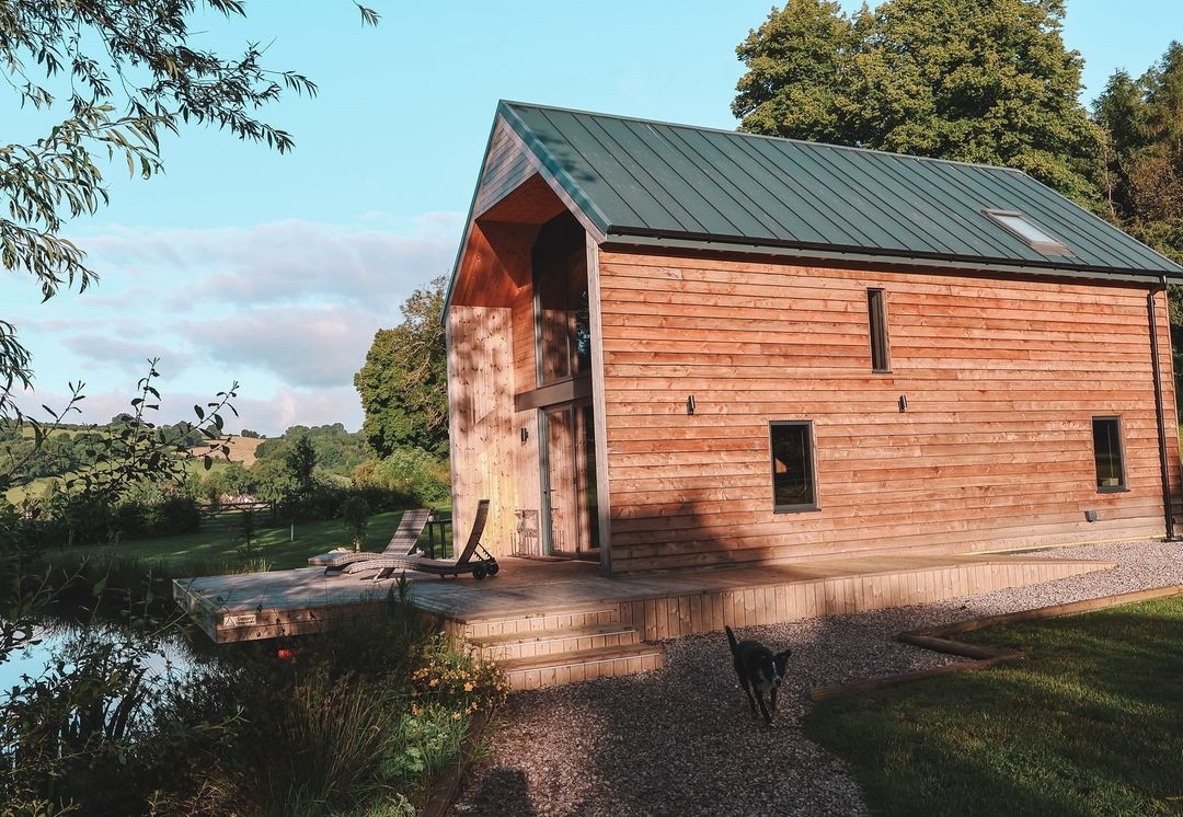 A cozy wooden cabin with a sloped green roof is situated by a tranquil pond, offering a hint of waterfront dining ambiance. It features large windows, a deck with chairs, and a dog on the gravel path. Surrounding the cabin are lush trees and grassy areas under a clear blue sky.