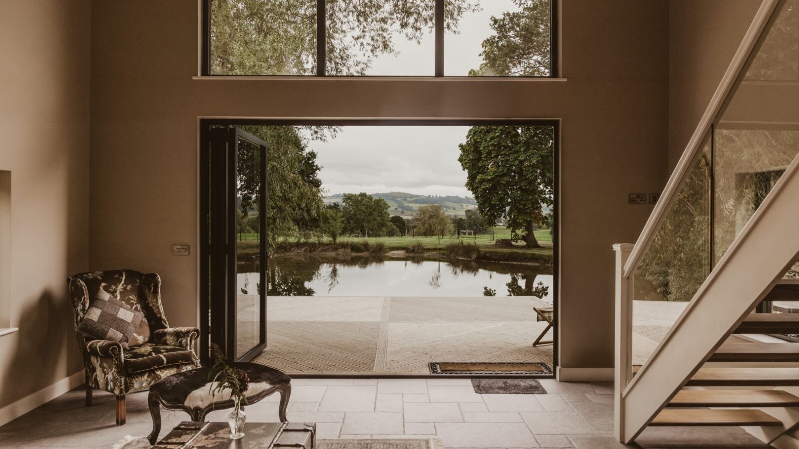 Spacious room with open French doors leading to a patio, overlooking a tranquil pond and lush greenery, perfect for waterfront dining. Stairs on the right.